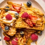 A close up of a plate of protein french toast topped with fresh strawberries, fresh blueberries, fresh raspberries, a dollop of greek yogurt, peanut butter, and maple syrup.