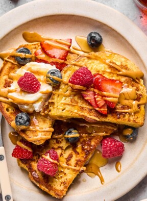 A close up of a plate of protein french toast topped with fresh strawberries, fresh blueberries, fresh raspberries, a dollop of greek yogurt, peanut butter, and maple syrup.