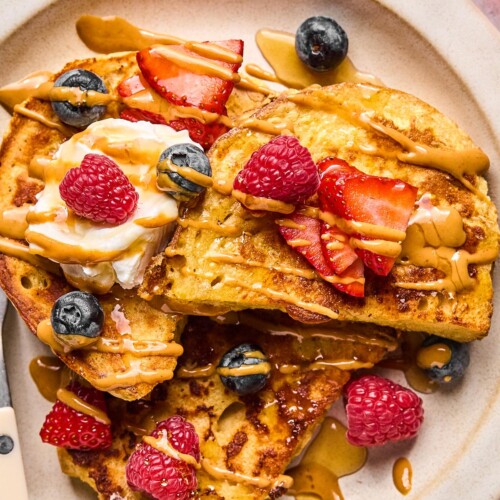 A close up of a plate of protein french toast topped with fresh strawberries, fresh blueberries, fresh raspberries, a dollop of greek yogurt, peanut butter, and maple syrup.