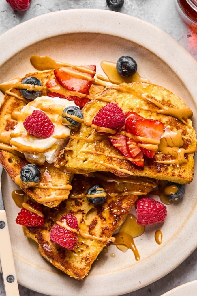 A close up of a plate of protein french toast topped with fresh strawberries, fresh blueberries, fresh raspberries, a dollop of greek yogurt, peanut butter, and maple syrup.