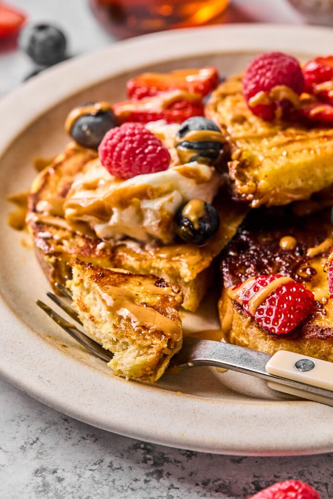 Slices of high protein french toast on a plate with a bite-size piece on a fork in the forefront. The french toast is topped with fresh berries, greek yogurt, peanut butter, and maple syrup.