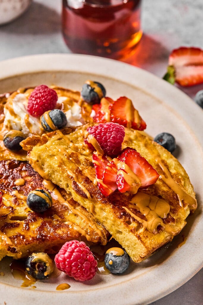 Pieces of healthy french toast topped with fresh berries, peanut butter, and maple syrup. Behind the plate is a jar of maple syrup and a strawberry that has been cut in half.