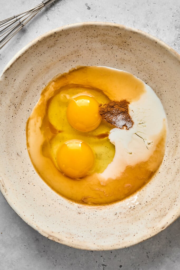Bowl filled with 2 cracked eggs, milk, cinnamon, and salt before it is whisked together. Next to the bowl is a metal whisk.
