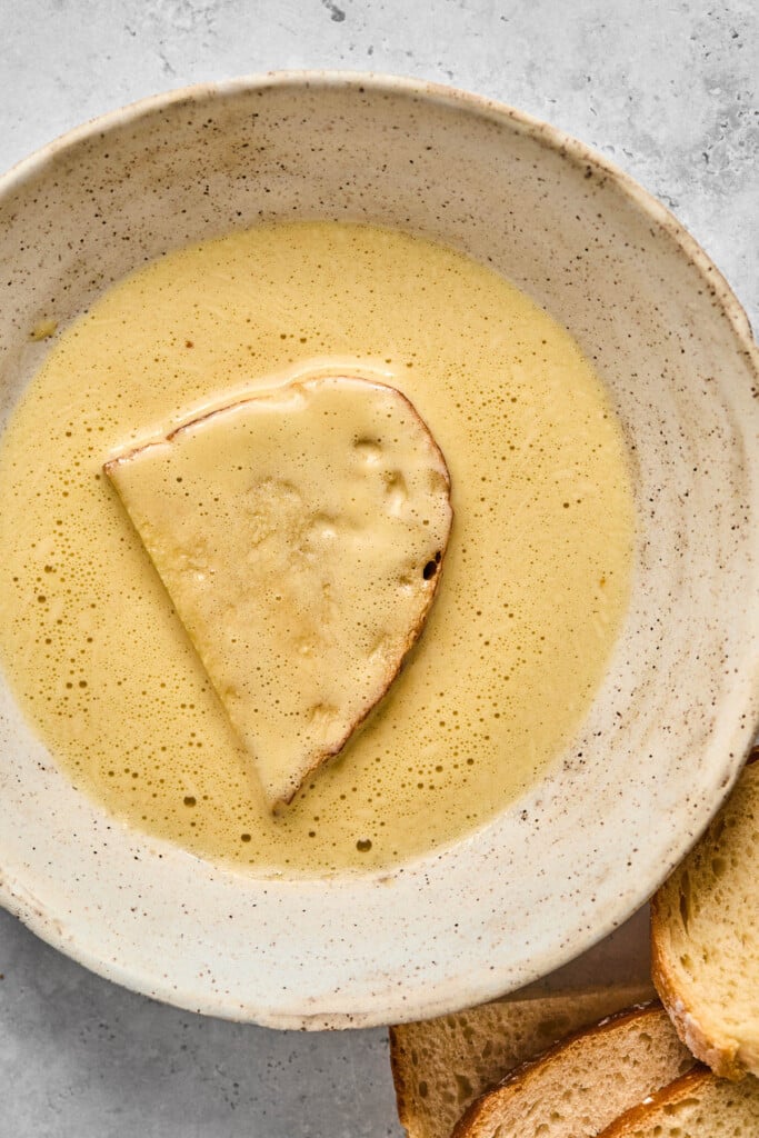 A white bowl with a piece of sliced bread in an egg mixture in it. Next to the bowl is more sliced bread.