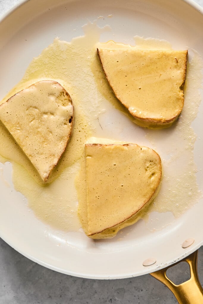 Three pieces of french toast cooking in a white skillet before they are flipped.