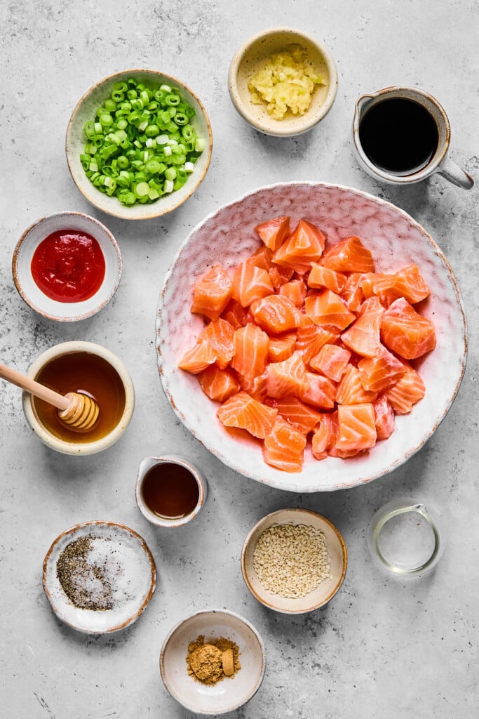 Grey concrete counter with a small bowl of minced garlic, a small cup of coconut aminos, a bowl of cubed raw salmon, a small cup of vinegar, a small bowl of sesame seeds, a small bowl of ground ginger, a small of bowl of salt and pepper, a small cup of sesame oil, a small dish of honey with a honey dipper in it, a small bowl of sriracha, and a bowl of diced green onions.