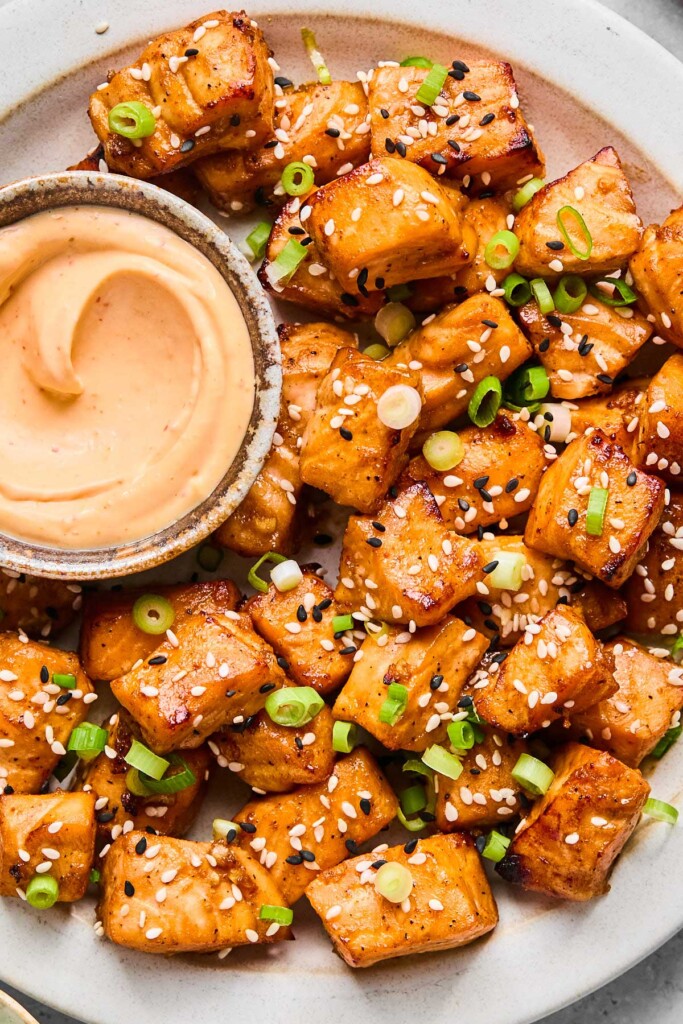 Overhead shot of a plate of air fryer salmon bites topped with diced green onions and sesame seeds. A dish of sriracha mayo is also on the plate.