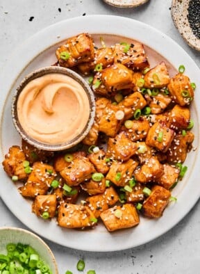 A plate of air fry salmon bites topped with diced green onions and sesame seeds. Also on the plate is a bowl of sriracha mayo for dipping. Around the plate is a bowl or more diced green onions and two different bowls of sesame seeds.
