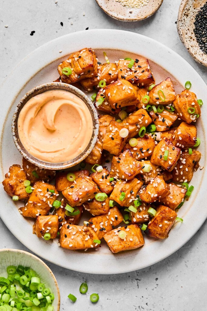 A plate of air fry salmon bites topped with diced green onions and sesame seeds. Also on the plate is a bowl of sriracha mayo for dipping. Around the plate is a bowl or more diced green onions and two different bowls of sesame seeds.