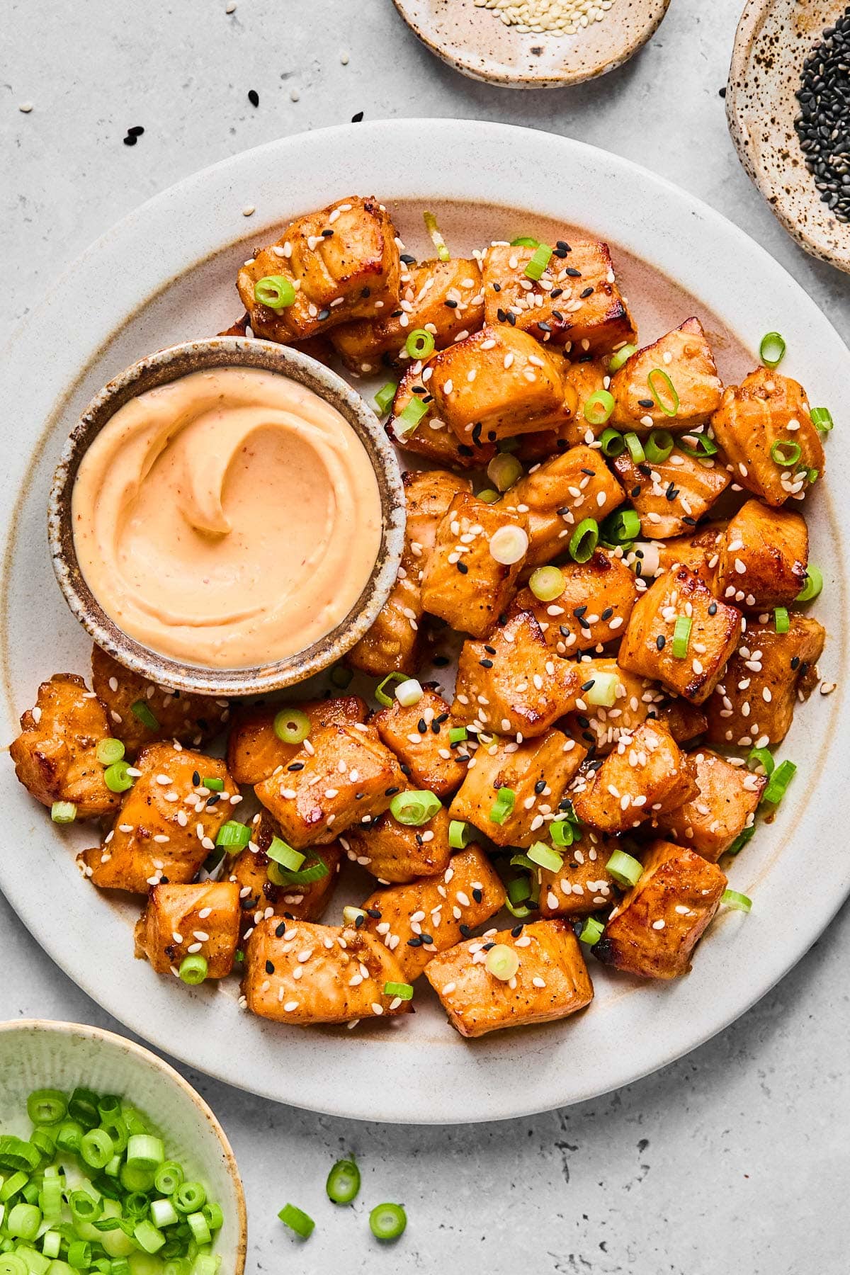 A plate of air fry salmon bites topped with diced green onions and sesame seeds. Also on the plate is a bowl of sriracha mayo for dipping. Around the plate is a bowl or more diced green onions and two different bowls of sesame seeds.