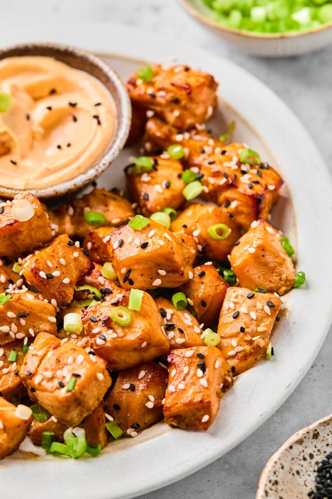 A close up of salmon air fryer bites on a white plate garnished with diced green onions and sesame seeds. Also on the plate is a small dish of sriracha mayo. Behind the plate is a bowl of diced green onions.