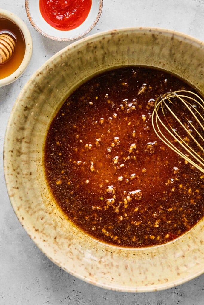 Bowl filled with a brown marinade with a whisk in it. Next to it is a small bowl of sriracha and a small bowl of honey.