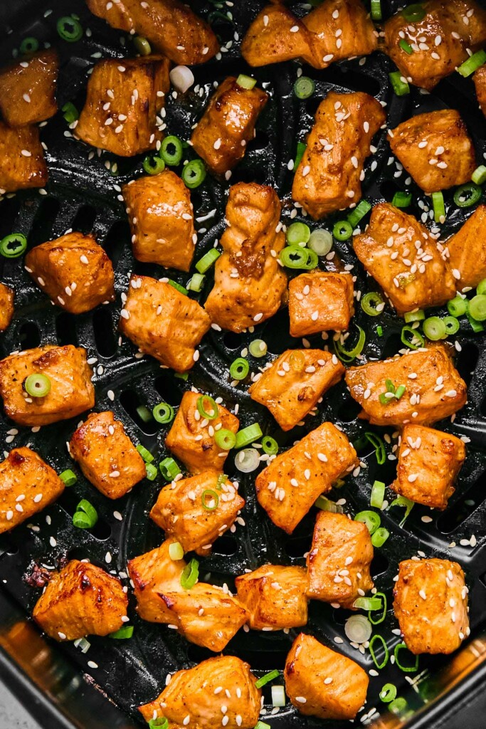 Salmon bites in an air fryer basket topped with sesame seeds and diced green onions.