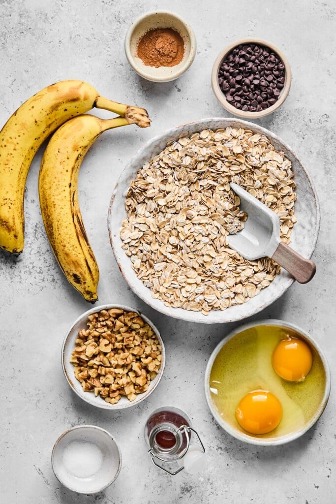 Grey concrete counter with a small bowl of cinnamon, a bowl of mini chocolate chips, a bowl of rolled oats with a scoop in it, two eggs that have been cracked into a small dish, a jar of maple syrup, a small dish of salt, a bowl of chopped walnuts, and two spotted bananas.