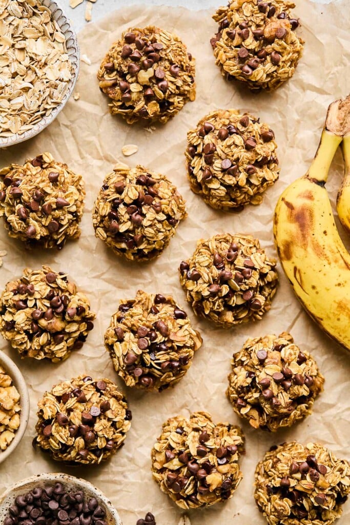 A bunch of oatmeal banana cookies spread across a piece of brown parchment paper. Around them is a spotted banana, a bowl of mini chocolate chips, a bowl of walnuts, and a bowl of rolled oats.