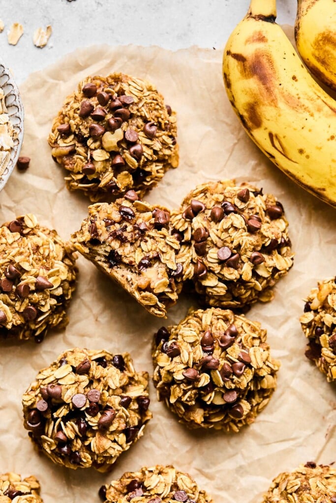 Overhead shot of banana oatmeal chocolate chip cookies on a piece of brown parchment paper. One of the cookies is standing on it's side and has a bite taken out of it. Next to the cookies are two spotted bananas.