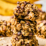 Three banana oatmeal cookies stacked on top of each other. The top one has a bite taken out of it. Around them are more cookies and in the far distance are some bananas.