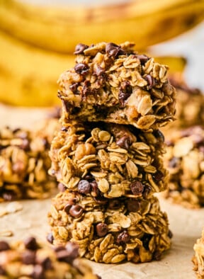 Three banana oatmeal cookies stacked on top of each other. The top one has a bite taken out of it. Around them are more cookies and in the far distance are some bananas.
