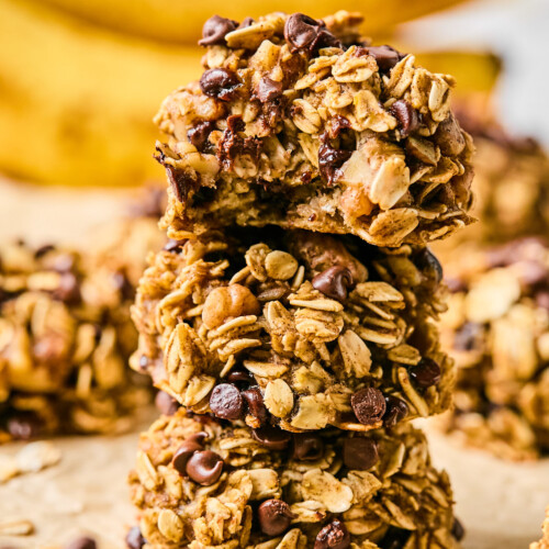 Three banana oatmeal cookies stacked on top of each other. The top one has a bite taken out of it. Around them are more cookies and in the far distance are some bananas.