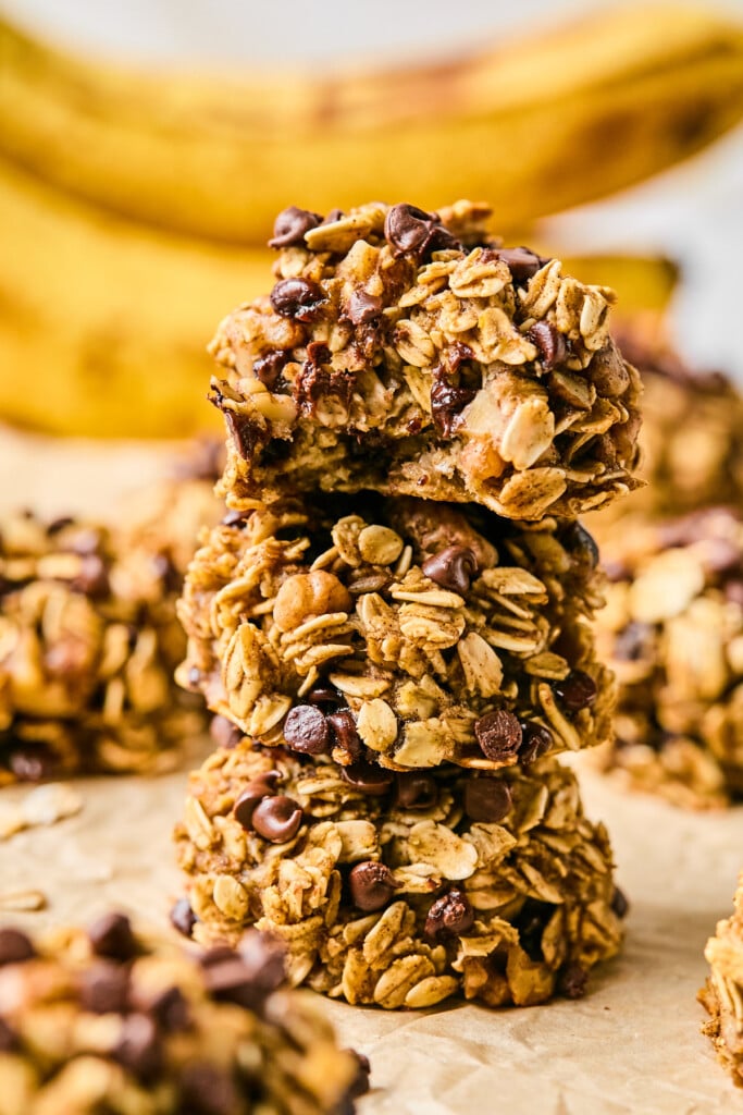 Three banana oatmeal cookies stacked on top of each other. The top one has a bite taken out of it. Around them are more cookies and in the far distance are some bananas.