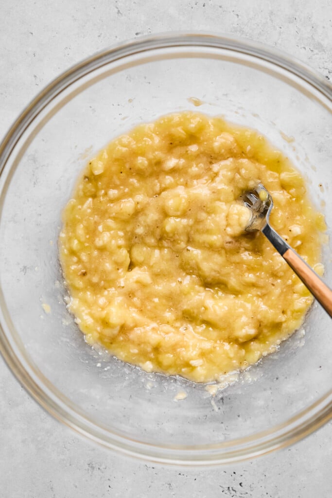 An overhead shot of a glass bowl with mashed banana in it. There is a fork in there as well.