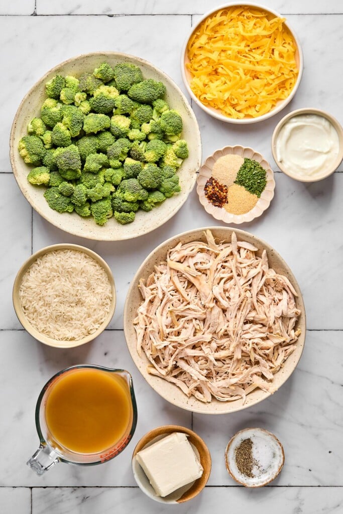 White tile counter with a bowl of orange shredded cheese, a bowl of greek yogurt, a bowl of spices, a bowl of shredded chicken, a bowl of salt and pepper, a small dish of cream cheese, a measuring cup of bone broth, a bowl of uncooked white rice, and a bowl of broccoli florets.