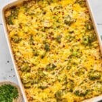Healthy chicken broccoli rice casserole covered with melted cheese and topped with red pepper flakes and fresh parsley in a large baking dish. Next to the dish is a small bowl of red pepper flakes, a small bowl of parsley, and a bowl of shredded cheese.