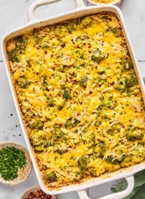 Healthy chicken broccoli rice casserole covered with melted cheese and topped with red pepper flakes and fresh parsley in a large baking dish. Next to the dish is a small bowl of red pepper flakes, a small bowl of parsley, and a bowl of shredded cheese.