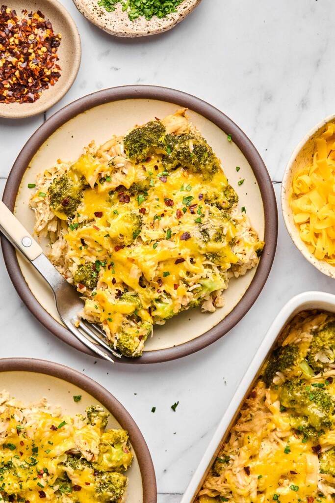 A piece of chicken broccoli and rice casserole with a fork next to it on a small plate. Around the plate is the dish of the rest of the casserole, another small plate with a piece of casserole on it, a small dish of red pepper flakes, and a small dish of fresh parsley.