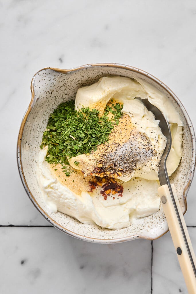 Bowl filled with greek yogurt, cream cheese, spices, red pepper flakes, salt, and pepper before it is mixed together. A spoon is also in the bowl.