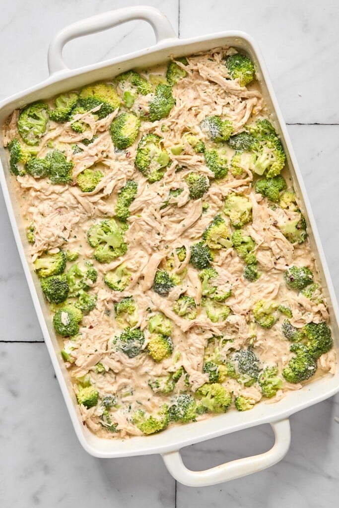 Overhead shot of a large white casserole dish filled with a mixture of cream cheese, broccoli, shredded chicken, and broth before it is baked.