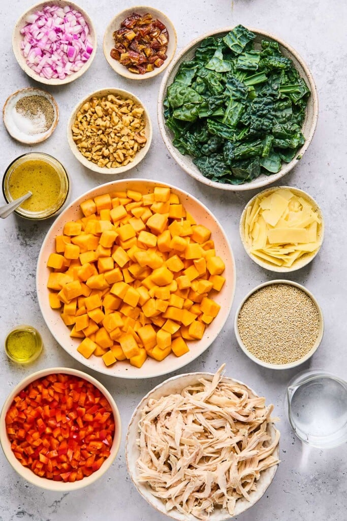 Grey concrete counter with a bowl of chopped kale, a bowl of shaved parmesan cheese, a bowl of uncooked quinoa, a measuring cup of water, a bowl of shredded chicken, a bowl of diced red bell pepper, a small dish of olive oil, a bowl of raw diced butternut squash, a jar of salad dressing with a whisk in it, a bowl of walnuts, a small dish of salt and pepper, a bowl of chopped dates, and a bowl of diced red onion.