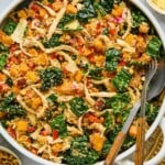 Chicken quinoa salad with butternut squash, kale, and vegges in a large white bowl. There is a fork and spoon in the bowl as well. Around the bowl is a a gold and white kitchen towel, a bowl of walnuts, a salt and pepper shaker, a jar of dressing, and a bowl of shaved parmesan cheese.