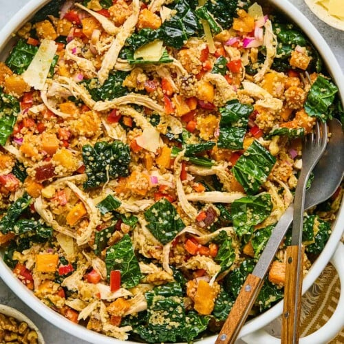 Chicken quinoa salad with butternut squash, kale, and vegges in a large white bowl. There is a fork and spoon in the bowl as well. Around the bowl is a a gold and white kitchen towel, a bowl of walnuts, a salt and pepper shaker, a jar of dressing, and a bowl of shaved parmesan cheese.