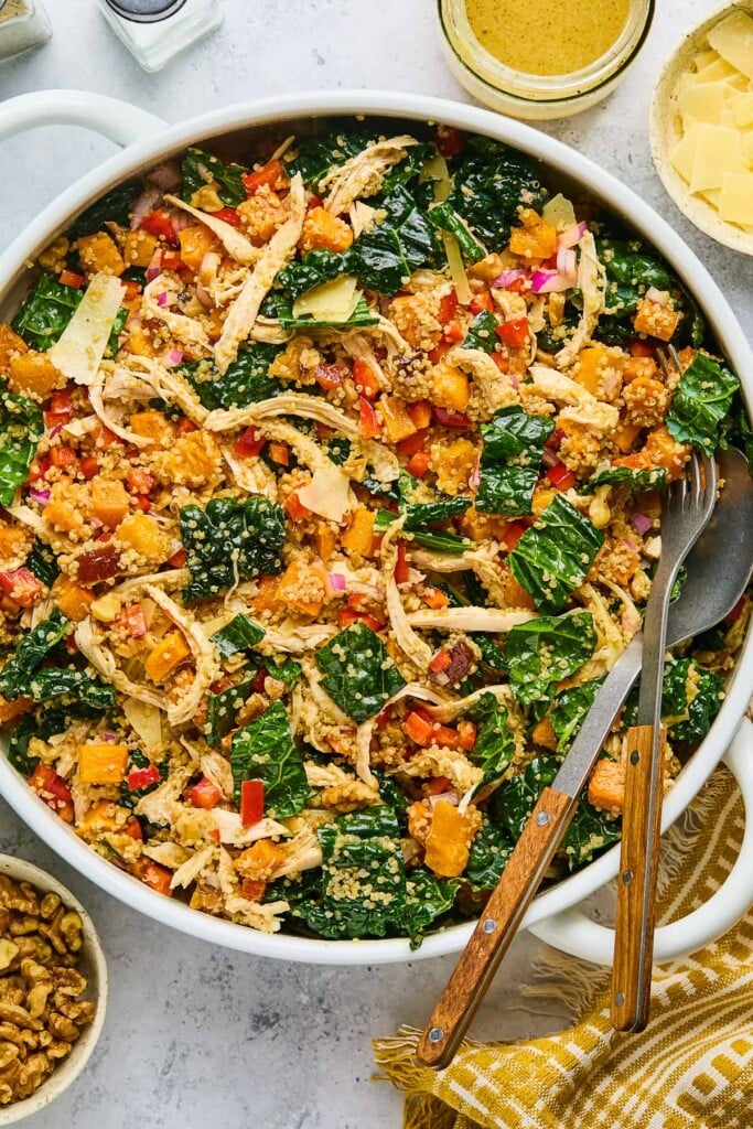 Chicken quinoa salad with butternut squash, kale, and vegges in a large white bowl. There is a fork and spoon in the bowl as well. Around the bowl is a a gold and white kitchen towel, a bowl of walnuts, a salt and pepper shaker, a jar of dressing, and a bowl of shaved parmesan cheese.
