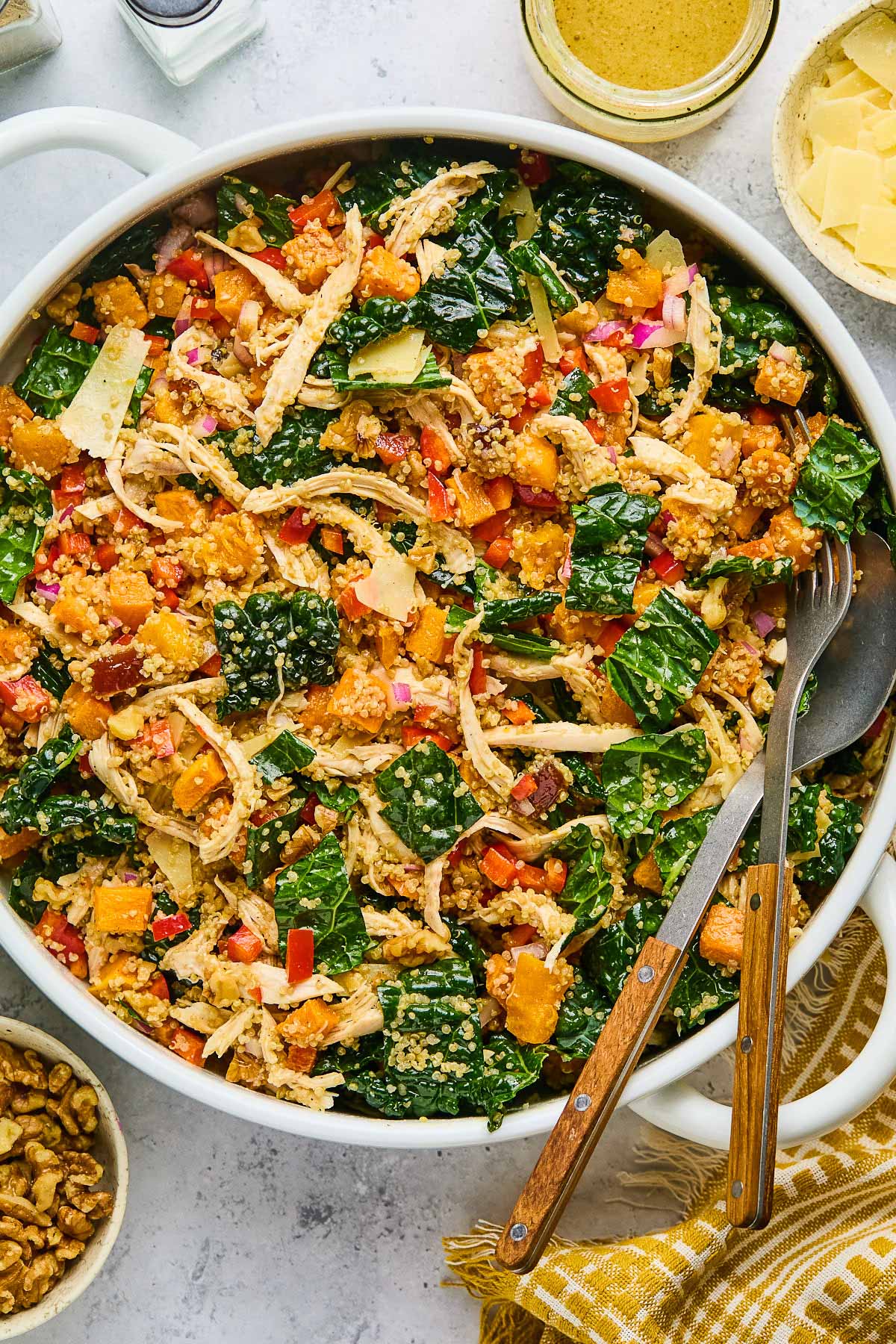 Chicken quinoa salad with butternut squash, kale, and vegges in a large white bowl. There is a fork and spoon in the bowl as well. Around the bowl is a a gold and white kitchen towel, a bowl of walnuts, a salt and pepper shaker, a jar of dressing, and a bowl of shaved parmesan cheese.