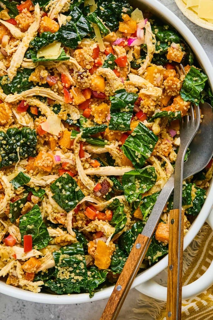Overhead shot of chicken and quinoa salad with kale and veggies in a large white bowl. A fork and spoon is also in the bowl. Around the bowl a tan and white kitchen towel.
