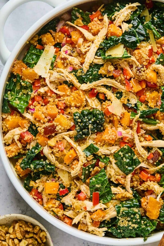Kale quinoa salad with shredded chicken, butternut squash, and veggies in a large white bowl. Next to the bowl is a small bowl of walnuts.