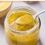 Mustard vinaigrette in a jar displaying a spoonful of dressing overtop the jar. Behind the jar is a bowl of mustard.