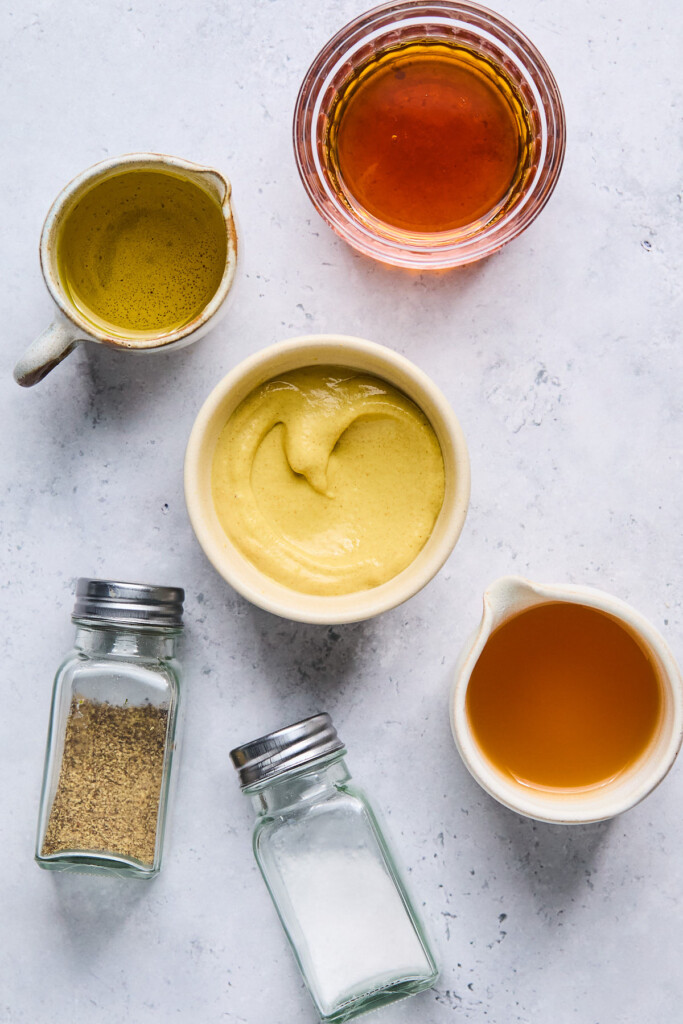 Grey concrete counter with a jar of maple syrup, a bowl of dijon mustard, a dish of apple cider vinegar, a salt shaker, a pepper shaker, and a measuring cup of olive oil.