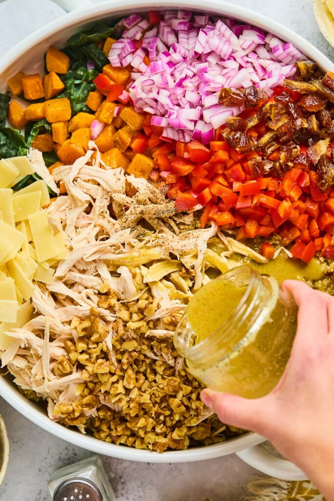 Large bowl with kale, diced red onion, chopped dates, diced red bell pepper, shredded chicken, walnuts, and parmesan cheese with dressing being poured overtop of it.