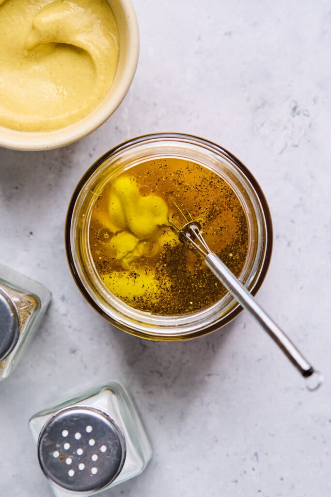 Overhead shot of a glass jar filled with olive oil, vinegar, dijon mustard, salt, and pepper. Around the jar is a bowl of mustard and salt and pepper shaker.
