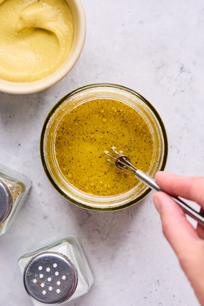 Overhead shot of someone whisking a jar of salad dressing. Around the jar is a bowl of mustard and salt and pepper shaker.
