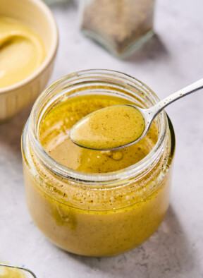 Mustard vinaigrette in a jar displaying a spoonful of dressing overtop the jar. In front of the jar is a small whisk and behind the jar is a bowl of mustard and a pepper shaker.