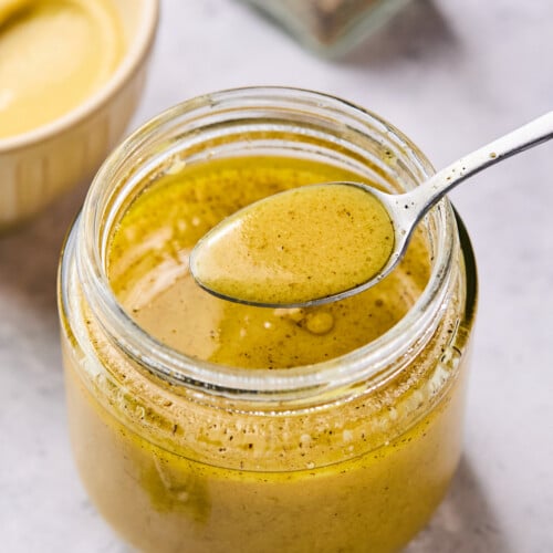 Mustard vinaigrette in a jar displaying a spoonful of dressing overtop the jar. In front of the jar is a small whisk and behind the jar is a bowl of mustard and a pepper shaker.