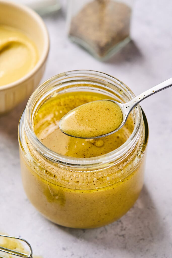 Mustard vinaigrette in a jar displaying a spoonful of dressing overtop the jar. In front of the jar is a small whisk and behind the jar is a bowl of mustard and a pepper shaker.