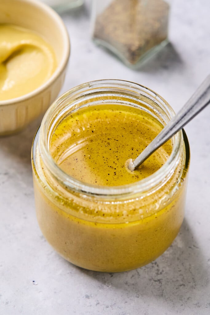 A jar of dijon mustard vinaigrette with a spoon in it. Behind the jar is a bowl of mustard and a pepper shaker.