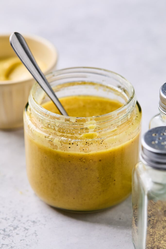 Maple mustard vinaigrette in a jar with a spoon in it. Around the jar is a pepper shaker and a bowl of mustard.