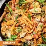 Large skillet filled with vegan pasta with veggies tossed in a creamy red sauce. It is garnished with fresh basil and grated cheese and there is a wooden spoon in the pan.