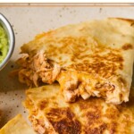 A plate with pieces of buffalo chicken quesadilla on it with a small dish of guacamole off to the side.